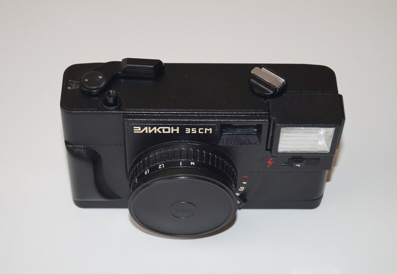 May include: Black Elikon 35CM camera with a lens cap. The camera has a flash and a silver-colored button on top. The text "ЭЛИКОН 35 CM" is printed on the front of the camera. The camera is on a white surface.