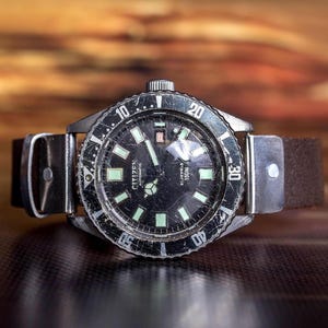 Puede incluir: Un reloj de buceo autom&aacute;tico Citizen vintage con esfera negra y marcadores luminosos. El reloj tiene un bisel giratorio y una correa de cuero marr&oacute;n. La esfera muestra el nombre de la marca e informaci&oacute;n de resistencia al agua.