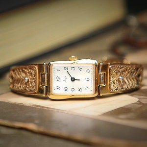 May include: A gold-toned vintage wristwatch with an engraved band and rectangular face. The watch face is white with black numerals and hands. The band features an ornate, floral-like design. The watch is displayed on a vintage photograph.