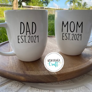 May include: Two white ceramic mugs with black text. The mug on the left says "Dad Est. 2021" and the mug on the right says "Mom Est. 2021". The mugs are sitting on a wooden coaster.