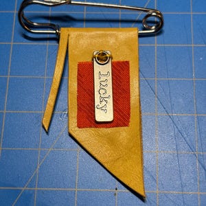 May include: A handmade pin featuring a yellow leather pennant with a red plaid square and a metal tag that says "Lucky". The pin is attached to a large gold safety pin.