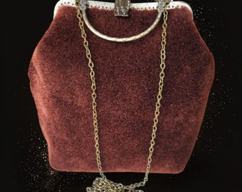May include: A burgundy velvet handbag with a gold chain strap. The clasp is ornate with a decorative metal frame. The bag has a rounded shape and a vintage aesthetic. The chain is a delicate, linked design.