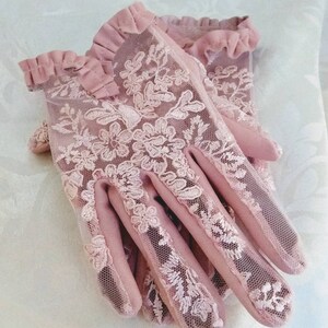 May include: A pair of pink lace gloves with a ruffled trim. The gloves are made of a delicate mesh fabric with floral embroidery.