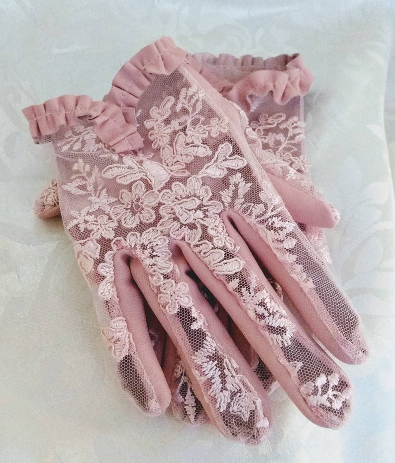 May include: A pair of pink lace gloves with a ruffled trim. The gloves are made of a delicate mesh fabric with floral embroidery.