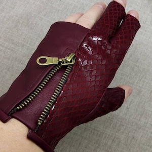 Pode incluir: Luvas sem dedos de couro bordô com padrão de pele de cobra e detalhe de zíper.