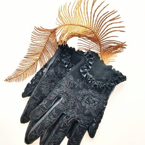 May include: A pair of black lace gloves with a ruffled trim. The gloves are lying on a white surface with a gold-coloured feather behind them.