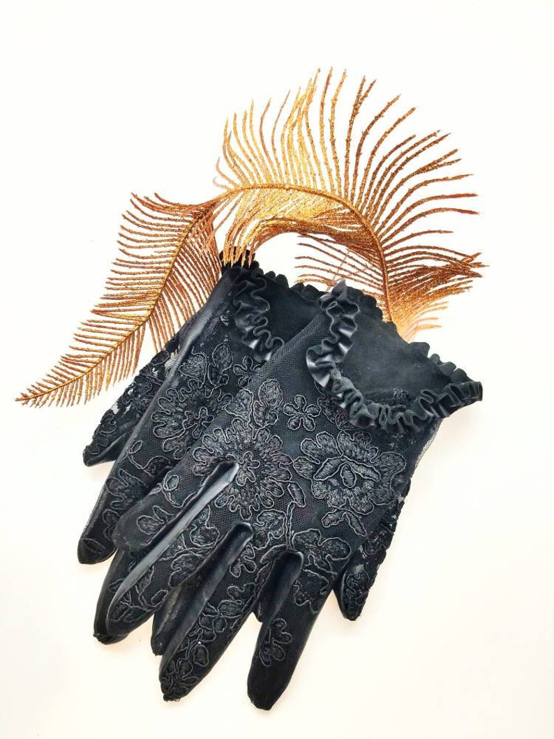 May include: A pair of black lace gloves with a ruffled trim. The gloves are lying on a white surface with a gold-coloured feather behind them.