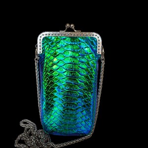Op de afbeelding: Een kleine, iriserende groene en blauwe clutch met een schubbenachtig patroon. De tas heeft een zilverkleurige metalen sluiting en een kettingriem. De achtergrond is zwart.