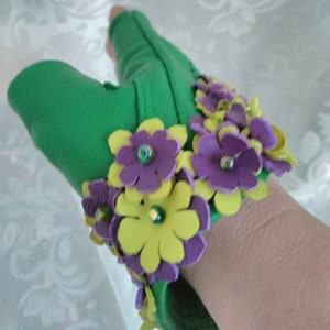 May include: Green leather fingerless gloves with a wrist cuff adorned with yellow and purple felt flowers. The flowers have a layered design and are accented with small, iridescent beads.