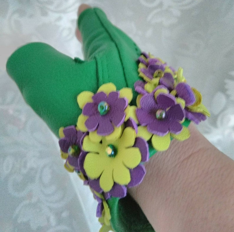 May include: Green leather fingerless gloves with a wrist cuff adorned with yellow and purple felt flowers. The flowers have a layered design and are accented with small, iridescent beads.