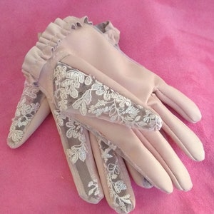 May include: A pair of light pink gloves with delicate white lace detailing. The gloves have a ruffled edge at the wrist.