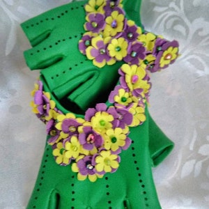 May include: Green fingerless gloves with purple and yellow flower embellishments. The flowers are made of felt and have a 3D effect.