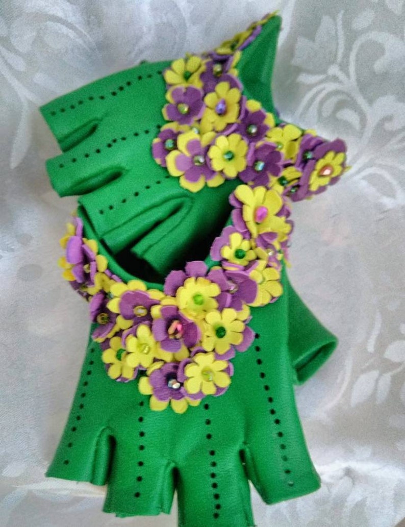 May include: Green fingerless gloves with purple and yellow flower embellishments. The flowers are made of felt and have a 3D effect.