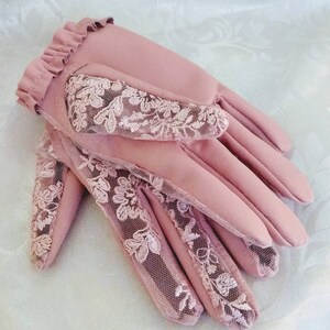 May include: A pair of pink leather gloves with delicate lace detailing. The gloves have a ruffled cuff and a floral lace pattern.