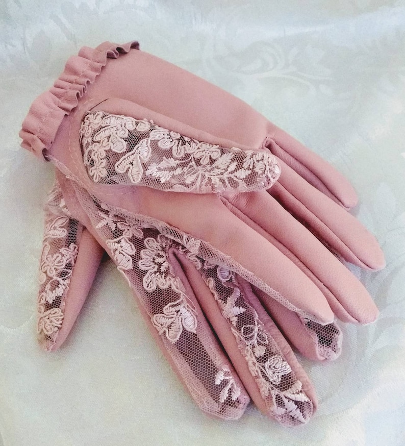 May include: A pair of pink leather gloves with delicate lace detailing. The gloves have a ruffled cuff and a floral lace pattern.