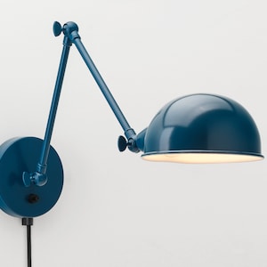 Può includere: Una lampada da parete blu navy con un braccio regolabile e un paralume a forma di semicerchio. La lampada è in metallo e ha una base rotonda. La lampada è collegata a una presa elettrica con un cavo nero.