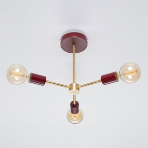 Puede incluir: Una moderna lámpara de techo con tres bombillas expuestas. La lámpara tiene un disco central y detalles cilíndricos de color burdeos, con brazos y portalámparas de color latón. Las bombillas son redondas y emiten una luz cálida.