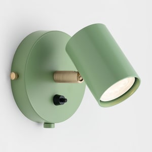 May include: A sage green wall-mounted spotlight with a cylindrical head and a circular base. The light fixture has a brass-coloured joint and a black on/off switch. The light is directed downwards, illuminating the area below.