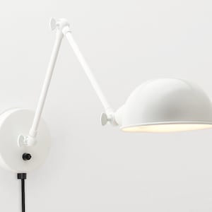 Könnte beinhalten: Verstellbare weiße Wandleuchte mit halbrundem Schirm. Die Lampe hat einen runden Sockel, einen beweglichen Arm und ein schwarzes Netzkabel. Sie ist für die Wandmontage konzipiert und bietet gezielte Beleuchtung.
