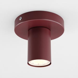 Puede incluir: Una lámpara de techo de color burdeos. La lámpara tiene un cuerpo cilíndrico y una base circular. Está montada en un techo blanco. La lámpara es de diseño moderno.