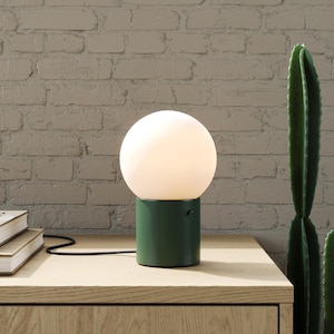 Könnte beinhalten: Eine moderne Tischlampe mit einem kugelförmigen, weißen Schirm auf einem zylindrischen, grünen Sockel. Die Lampe steht auf einer Holzoberfläche neben einem Stapel Bücher und einem Bleistift. Ein Kaktus ist im Hintergrund.