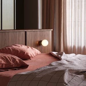 K&ouml;nnte beinhalten: Ein Bett mit einem Kopfteil aus dunklem Holz und einer kleinen runden Wandleuchte. Die Bettw&auml;sche besteht aus terrakottafarbenen Kissen und einem Laken sowie einer hellen Decke mit einem dunklen, gitterartigen Muster.