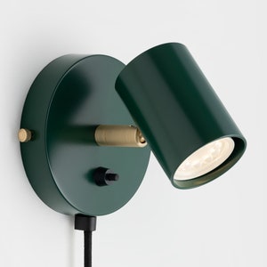 Op de afbeelding: Een groene wandlamp met een messing accent en een zwarte aan/uit-schakelaar. De lamp is aangesloten en heeft een zwart snoer.