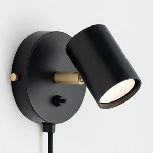 May include: A black wall-mounted spotlight with a brass accent and a cord. The light is on and the bulb is visible.