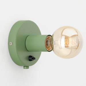 Op de afbeelding: Een groene wandlamp met een ronde basis en een cilindrische arm. De lamp heeft een zwarte aan/uit-schakelaar en is voorzien van een heldere glazen lamp.