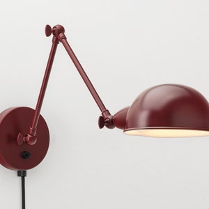 Op de afbeelding: Een bordeauxrode wandlamp met een verstelbare arm en een koepelvormige kap. De lamp is bevestigd aan een cirkelvormige basis met een schakelaar en een zwart netsnoer. Het ontwerp is minimalistisch en modern.