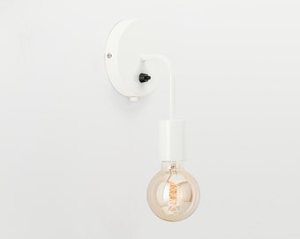 May include: A white wall-mounted light fixture with a curved arm and a cylindrical base. The fixture holds a large, round, Edison-style light bulb. The light bulb has a warm, amber glow. The fixture is mounted on a white wall.