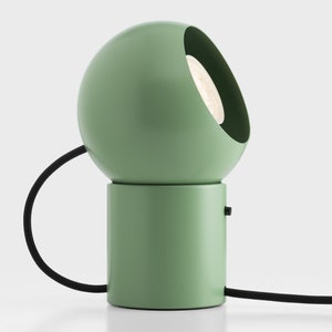 Może przedstawiać: Zielona lampa stołowa z okrągłym kloszem i cylindryczną podstawą. Lampa ma czarny przewód i białą żarówkę.