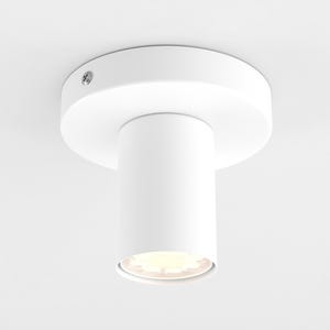 May include: A white, cylindrical ceiling light fixture with a circular base. The light fixture is mounted on a white ceiling and has a small screw visible on the base. The light is illuminated.