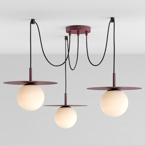 May include: A modern three-light pendant lamp with a burgundy finish. The fixture features three spherical, frosted glass globes suspended from the ceiling by black cords. Each globe is accented with a burgundy disc shade.