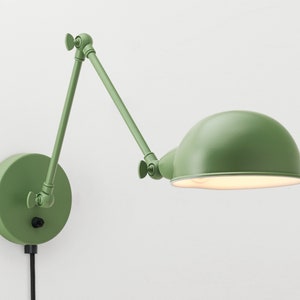 May include: A green wall-mounted adjustable lamp with a rounded shade. The lamp has a circular base and an articulated arm, allowing for directional lighting. The lamp is painted in a muted green color and has a black power cord.