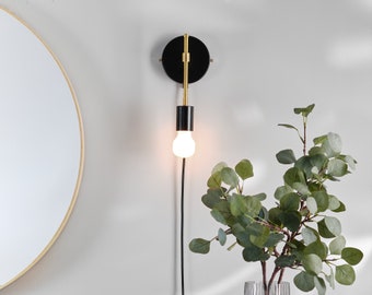 May include: A modern wall sconce with a black and gold finish. The sconce features a circular backplate, a vertical arm, and a black cylindrical lamp holder. A lit, round light bulb hangs from the holder. A black cord extends down. A round mirror and green plant are in the background.
