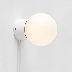 Op de afbeelding: Een witte wandlamp met een ronde, gloeiende lamp. De lamp is gemonteerd op een witte, cirkelvormige basis. Een witte kabel hangt naar beneden vanaf de armatuur. Modern design.