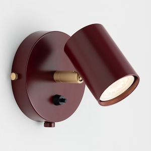 Op de afbeelding: Een rode wandspot met een draaibare messing arm en een zwarte aan/uit schakelaar. Het licht is naar beneden gericht.