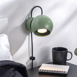 Può includere: Una lampada da parete verde con un paralume rotondo e un cavo nero. La lampada è montata su una parete bianca e si trova accanto a un comodino con una tazza nera e un quaderno.