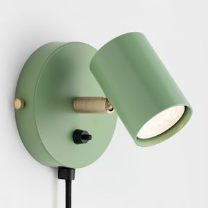 Op de afbeelding: Een moderne, saliegroene wandspot met een cilindrische kop en een cirkelvormige basis. De lamp heeft messing accenten en een zwarte aan/uit-schakelaar. Een zwart netsnoer loopt vanaf de basis.