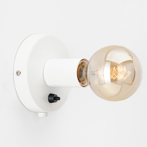 Op de afbeelding: Witte wandlamp met een ronde basis en een cilindrische arm. De wandlamp heeft een zwarte aan/uit-schakelaar en een heldere glazen lamp met een gouden afwerking.