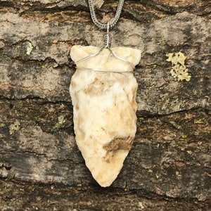 May include: A cream-colored arrowhead pendant with a silver chain. The arrowhead is wrapped with silver wire and has brown and tan markings. The background is a textured, dark brown surface.