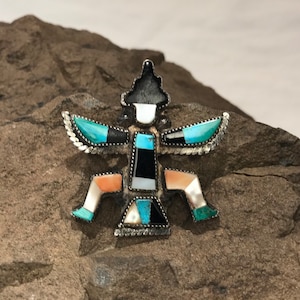 May include: A silver pendant with a geometric design inlaid with turquoise, black, white, and pink stones. The pendant is in the shape of a stylized human figure with outstretched arms.