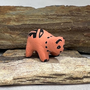 May include: A small, hand-painted ceramic bear figurine. The bear is pink with black markings and is standing on a wooden surface.