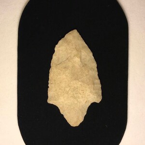 May include: A light brown arrowhead with a pointed tip and a slightly curved base. The arrowhead is displayed on a black velvet background.