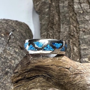 May include: A silver ring with a geometric design. The ring features a band of inlaid turquoise-colored stones set in a triangular pattern. The ring is displayed on a piece of weathered wood, showcasing its texture and color.