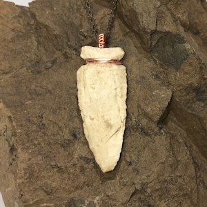 May include: A white stone arrowhead pendant with copper wire wrapping, hanging from a silver chain on a brown rock background.