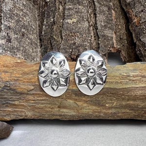 May include: A pair of silver-colored oval stud earrings. Each earring features a raised floral design with a central sphere and surrounding petal shapes. The earrings are displayed on a piece of wood.