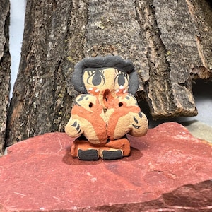May include: A small, handcrafted ceramic figurine with a stylized human form. The figure features a black head covering, large eyes, and orange and tan body markings. The figurine is set on a red stone surface, with a textured wood background.