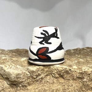 May include: A small, white ceramic thimble with black and red painted designs. The thimble features a black animal figure and red leaf-like shapes against a white background. The base and rim are black.